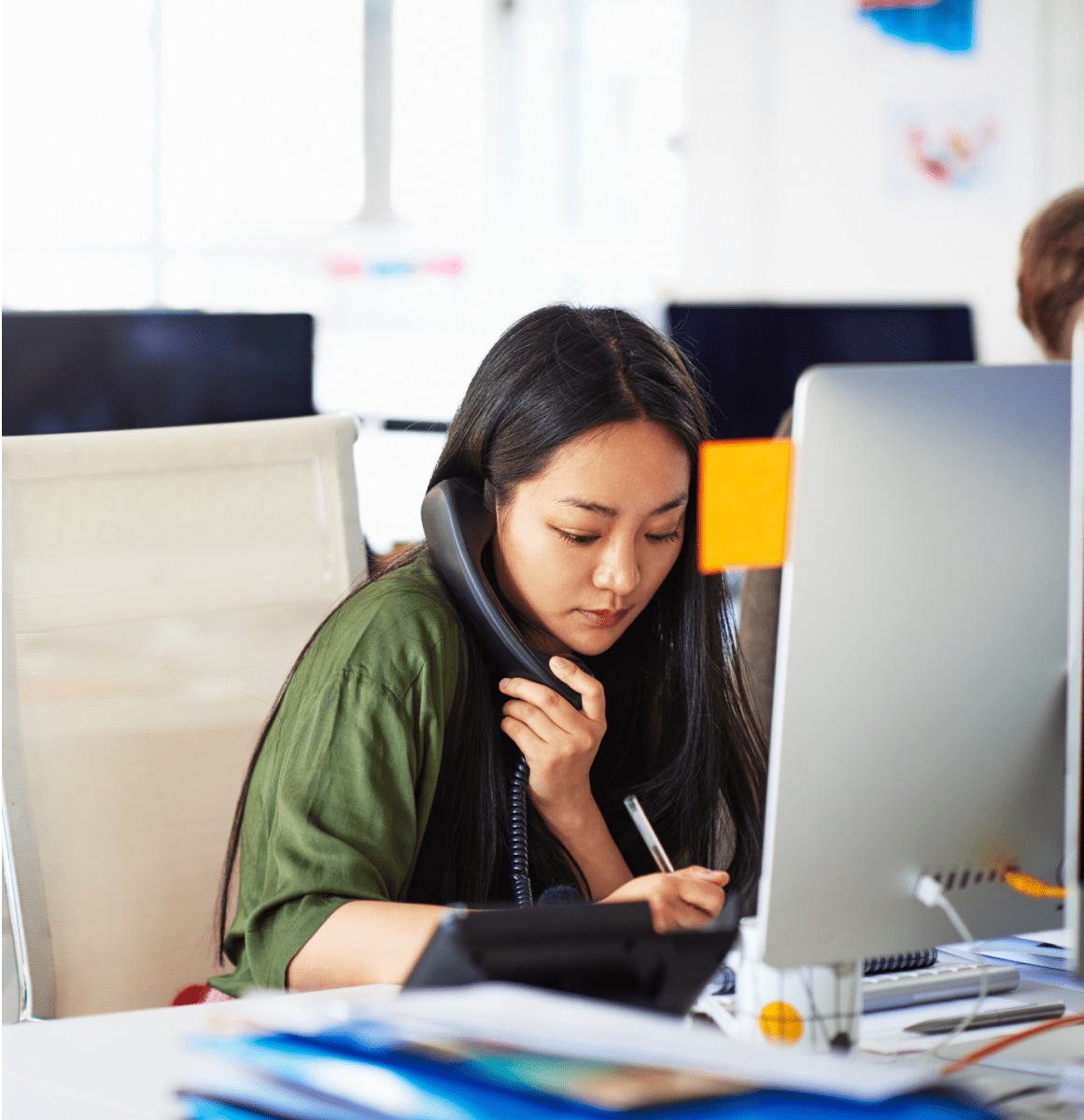 Professional on a phone call while taking notes at her desk in a modern office, representing media and press inquiries.